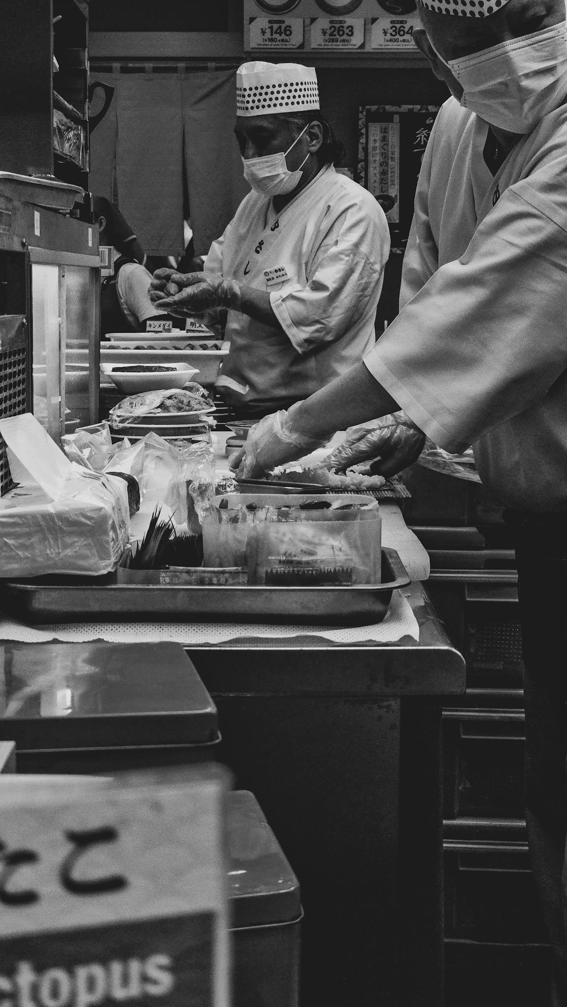Monochrome image of sushi chefs preparing dishes in a bustling Japanese kitchen.